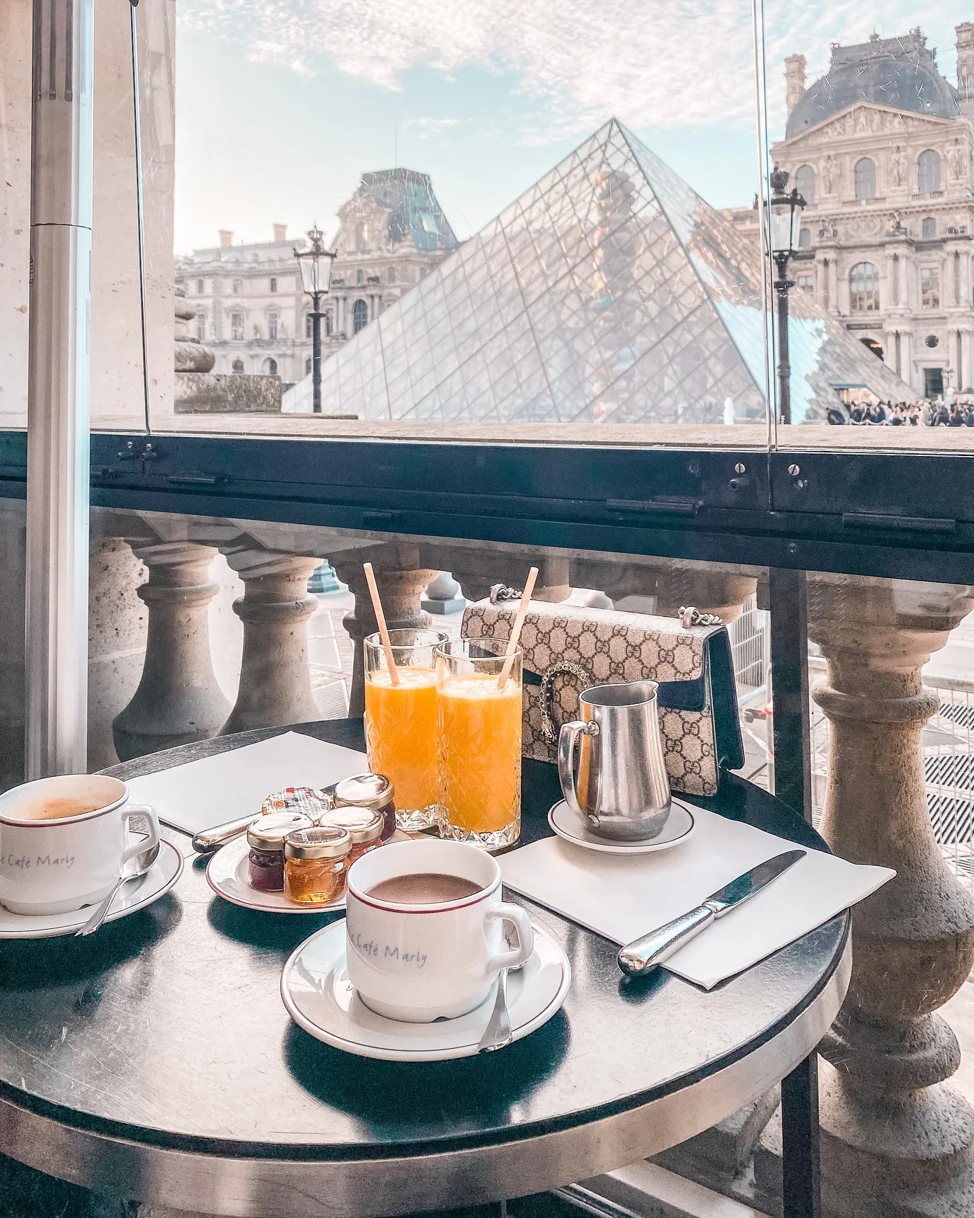 Table set with coffee, orange juice and condiments on a café balcony overlooking the Louvre Pyramid and historic museum façade.