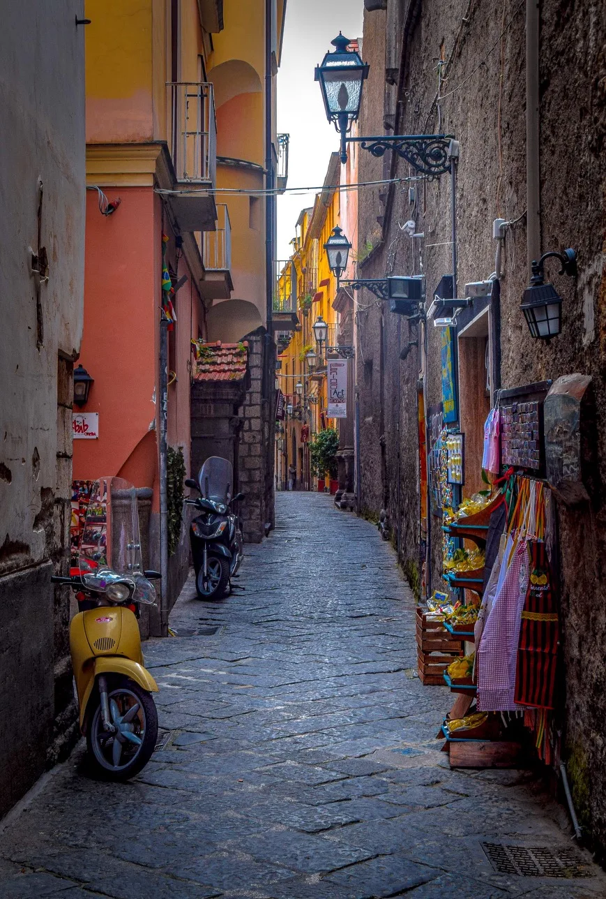 Narrow cobblestone alley lined with colourful Mediterranean buildings, scooters and small street shops displaying souvenirs.