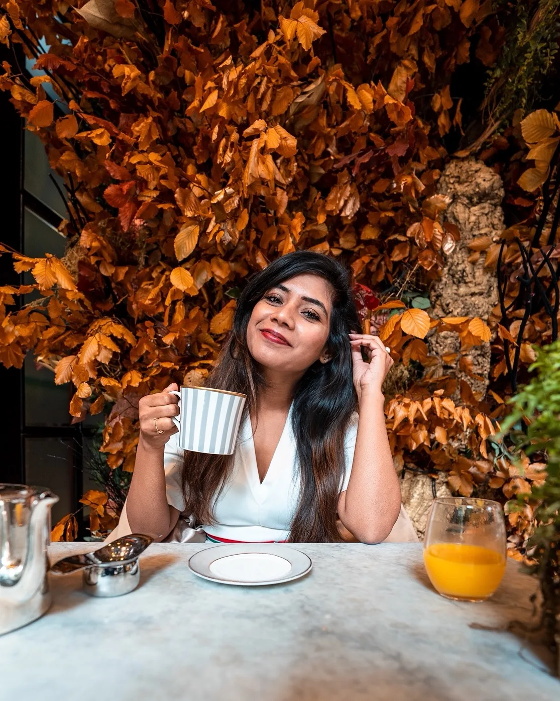 Woman smiles while holding a striped mug at a cafe table with orange autumn foliage behind her and a glass of orange juice nearby.