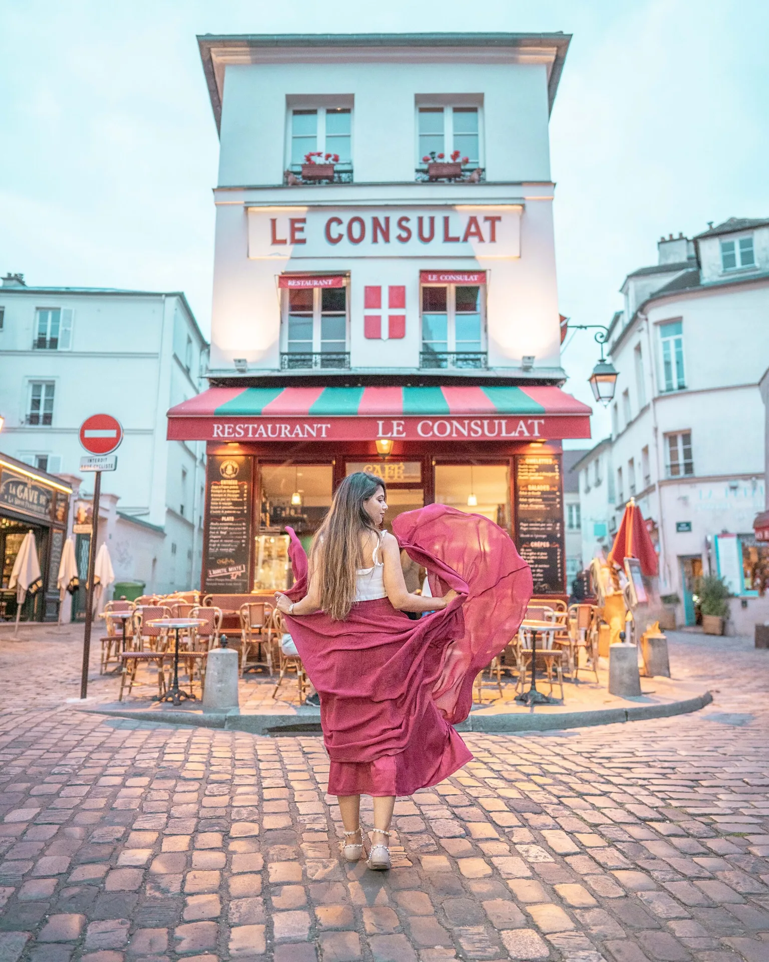 Woman in a flowing red skirt twirling on cobblestones in front of the Parisian café Le Consulat at dusk