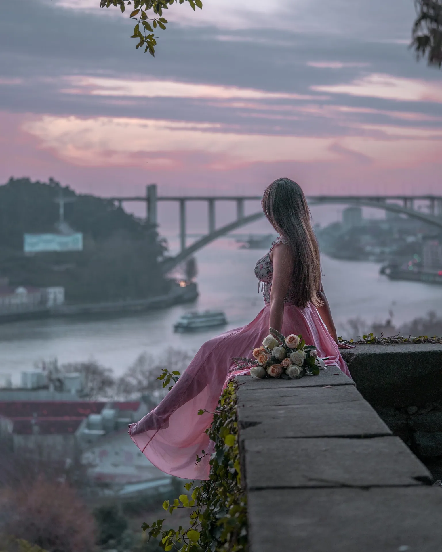 Woman in a flowing pink dress sits on a stone wall holding roses, overlooking a river and arched bridge at sunset.