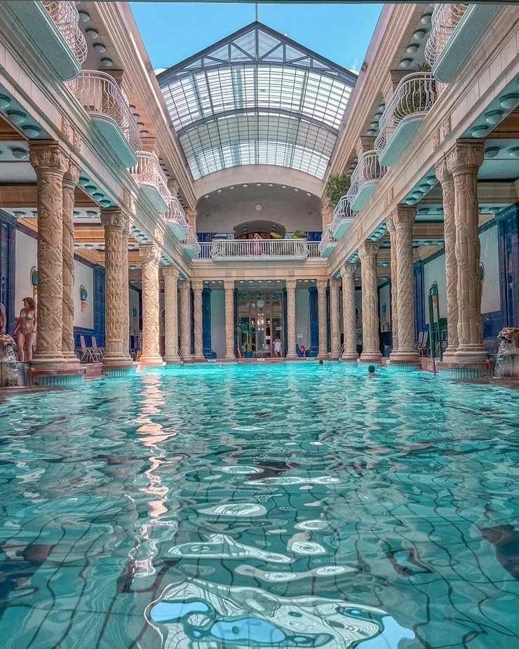 Indoor thermal pool with turquoise water framed by ornate carved columns and a glass vaulted ceiling in a historic bathhouse.