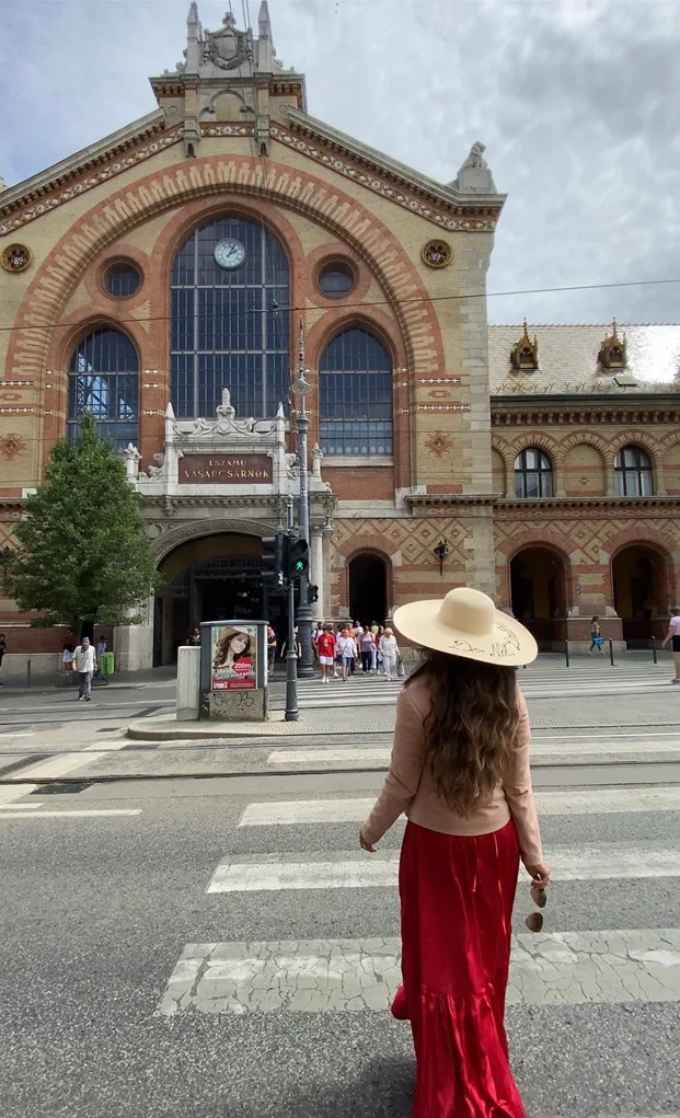 Woman in a wide-brimmed hat and red skirt crossing toward the ornate façade of Budapest's Great Market Hall.