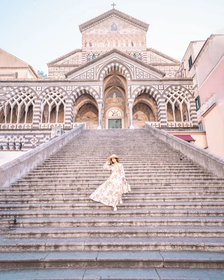Woman in a floral dress and wide-brim hat descending the wide stone steps in front of Amalfi Cathedral's striped Byzantine façade.