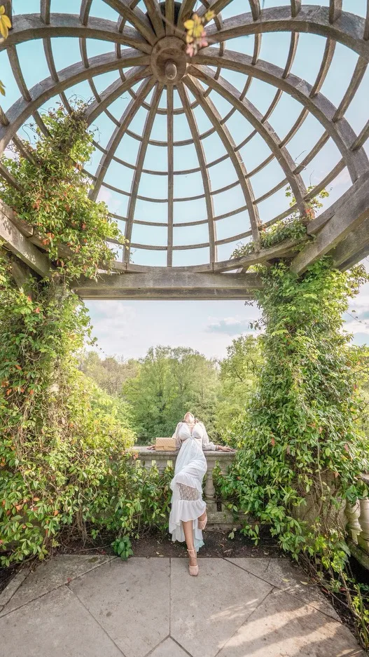 Woman in a flowing white dress leaning on a stone balustrade beneath a wooden domed pergola, overlooking a lush green park.