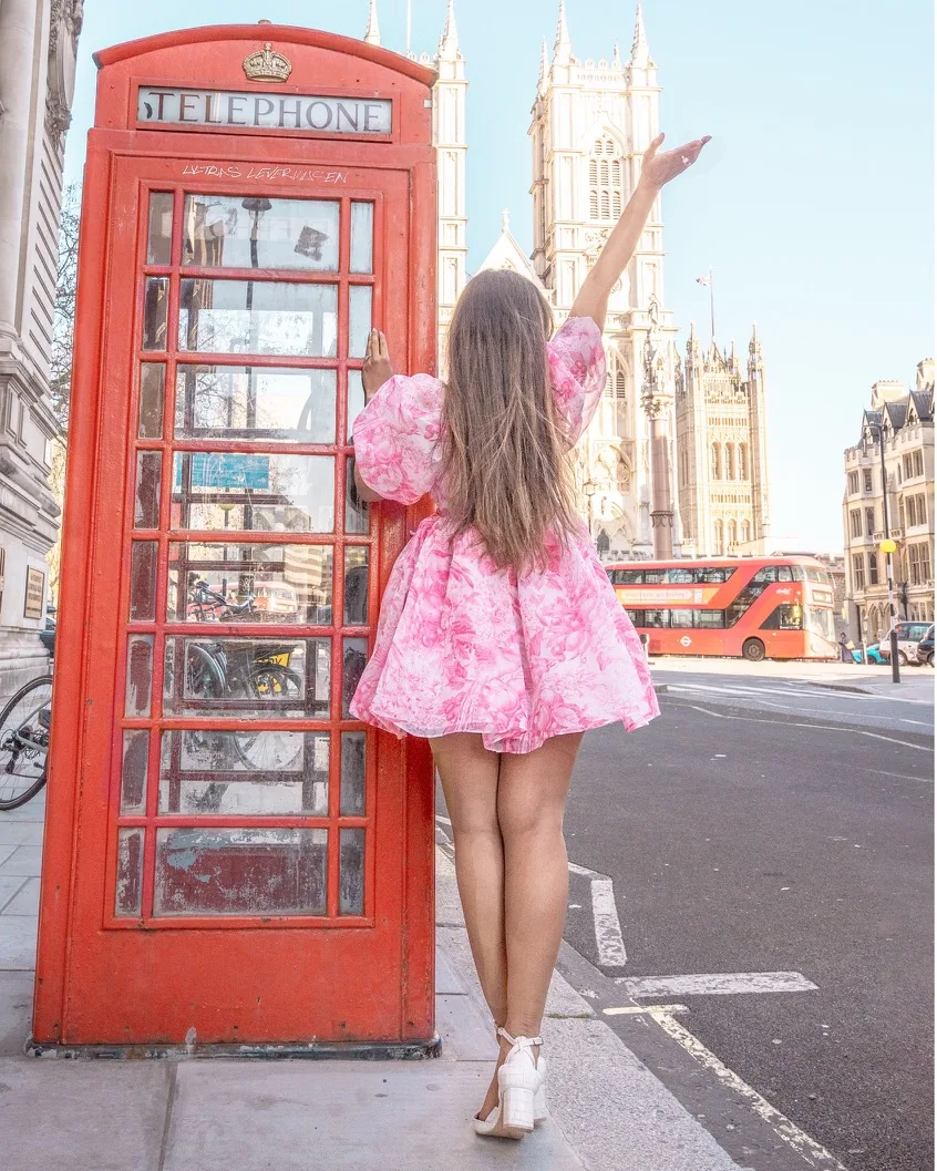 Woman in a pink dress posing with her arm raised beside a classic red London telephone box with Westminster Abbey and a red double-decker bus in the background.